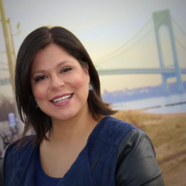 A smiling Jaclyn Tacaronte with dark hair stands outdoors near a body of water, with a large suspension bridge in the background on a cloudy day. She is wearing a blue top with black sleeves.
