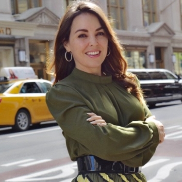 Nicole Cueto with long brown hair, wearing a green blouse and hoop earrings, smiles with arms crossed on a street in Manhattan, New York City. Yellow taxis and buildings are in the background.