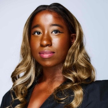 Shana Feggins with long, wavy, light brown hair and medium-dark skin looks confidently at the camera. She is wearing a black top and has subtle makeup with winged eyeliner, pink lipstick, and a neutral background.