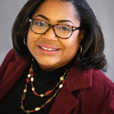 Yvette Richardson with shoulder-length dark hair, wearing glasses, a maroon blazer, a black top, and a multicolored beaded necklace, smiling at the camera against a plain background.