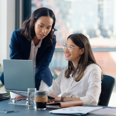 Two women in an office setting. One is standing and pointing at a laptop screen, while the other is seated, smiling and looking at the screen. There are notebooks and a coffee cup on the table. Large windows are in the background.