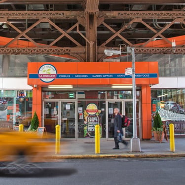 A bright orange storefront with a sign reading La Marqueta sits under a steel bridge. A blurred yellow taxi passes in the foreground, while a person in black walks by the entrance, which displays various signs and advertisements.