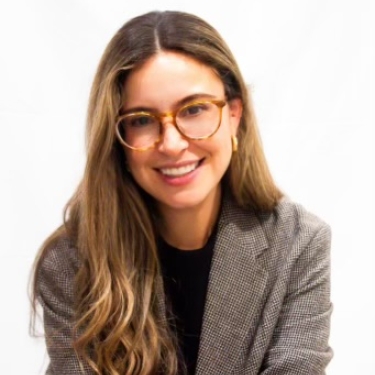 Cristina Garcia with long wavy brown hair, wearing glasses and a checkered blazer over a black top, smiles while sitting against a plain white background.