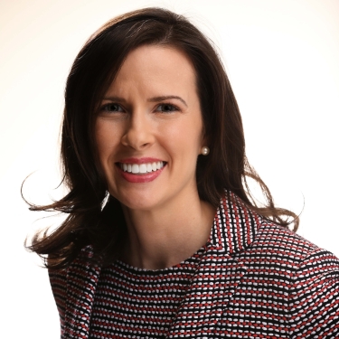 Kristan Lansbery with long brown hair smiles at the camera. She is wearing a red, black, and white patterned blazer over a black top, and the background is bright and white.