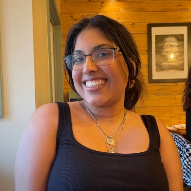 Shivanni Rambaran with long dark hair and glasses smiles while sitting in a cozy restaurant with wooden walls. She is wearing a black tank top and layered necklaces.