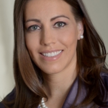 Susan Migliaccio with long brown hair, wearing a pearl necklace and earrings, and a shiny, purple blouse, smiling at the camera against a neutral background.