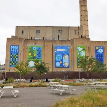 A large industrial building with colorful abstract murals on its exterior walls, depicting geometric shapes and patterns. There are trees, grass, and several white picnic tables in the foreground.