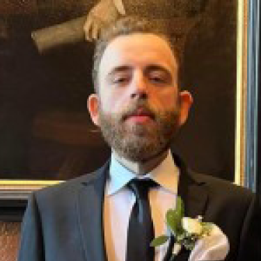 Allen Bogati with a beard and mustache is wearing a black suit, white shirt, and black tie, with a white boutonnière pinned to his lapel. He is standing indoors in front of a framed painting.