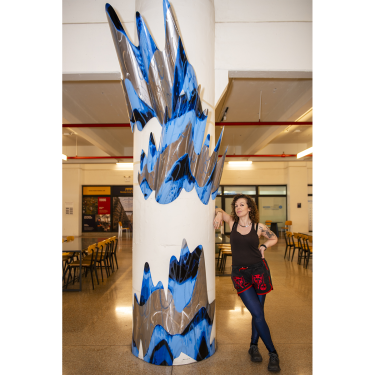 A woman stands beside a white column decorated with abstract, jagged blue and metallic shapes in a spacious room with tables and chairs in the background.
