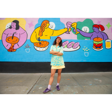 Hyesu Lee in colorful clothes stands smiling with arms crossed in front of a bright mural showing cartoon people eating and toasting with food and drinks. The mural features bold, cheerful colors and playful designs.