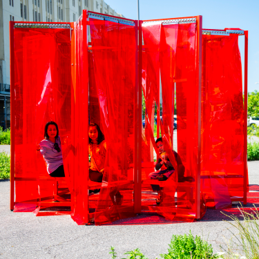 Three children sit inside a bright red, semi-transparent outdoor art structure made of panels, set on a sunny day at Brooklyn Army Terminal, with greenery and buildings visible in the background.