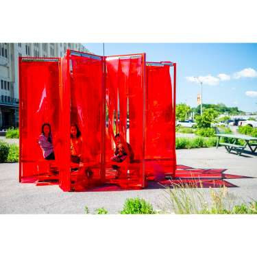 Three children sit inside a bright red, semi-transparent outdoor art structure made of panels, set on a sunny day at Brooklyn Army Terminal, with greenery and buildings visible in the background.