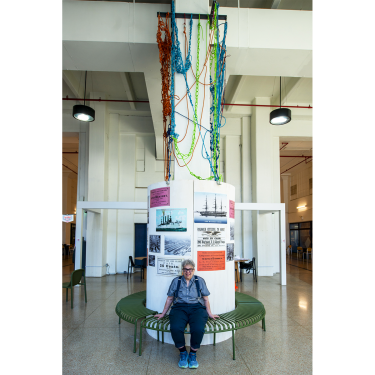 A person sits on a curved green bench in front of a cylindrical display with photos, text, and colorful ropes hanging from above in a spacious, modern indoor area with large columns and hanging lights.