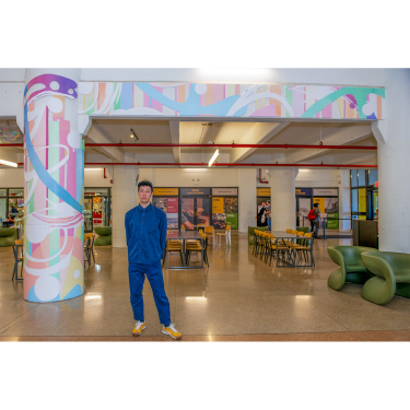 A person in a blue outfit stands in a spacious, modern indoor area with colorful abstract art on a column and wall. Tables, chairs, and people are visible in the background.