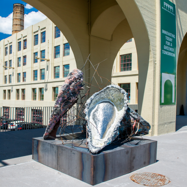 Large abstract sculptures resembling seashells and organic forms sit on a metal base beneath an archway outside Brooklyn Army Terminal building. A sign nearby reads “INNOVATING TODAY FOR A GREENER TOMORROW.”