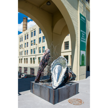 Large abstract sculptures resembling seashells and organic forms sit on a metal base beneath an archway outside Brooklyn Army Terminal building. A sign nearby reads “INNOVATING TODAY FOR A GREENER TOMORROW.”