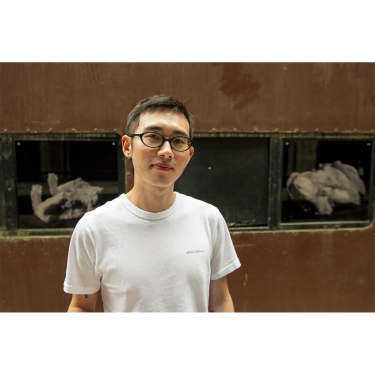 A person wearing glasses and a white t-shirt stands in front of a brown wall with glass windows displaying rock-like objects. The person is looking at the camera and smiling slightly.