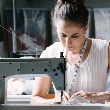 A woman wearing a white top and a measuring tape around her neck works intently at a sewing machine in a workshop, surrounded by fabric and storage bins in the background.