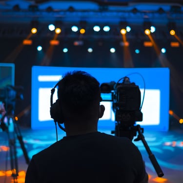 Silhouetted camera operators film in a studio with bright blue screens and stage lighting, capturing a production or live event. Multiple lights shine overhead, creating a dramatic atmosphere.