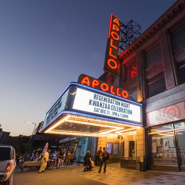 People walk past the illuminated Apollo Theater at dusk. The marquee advertises “Regeneration Night Kwanzaa Celebration, Sat Dec 31, 7PM &amp; 9:30PM.” The building’s neon Apollo sign stands out against the evening sky.