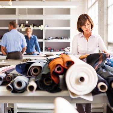 A woman examines fabric samples on a table covered with large rolls of cloth in a bright workspace; two people stand talking in the background near shelves filled with more fabric.