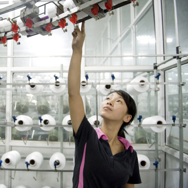 A woman in a black and pink shirt reaches up to adjust machinery in a textile factory, with rows of large white spools of thread in the background.