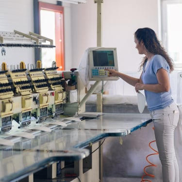 A woman operates a digital control panel next to a row of embroidery machines in a modern textile workshop. She is holding fabric, wearing a light blue shirt and jeans, and standing in a bright, clean workspace.