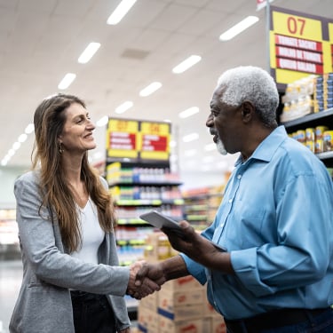 A woman and a man shake hands and smile at each other in a grocery store aisle with shelves of canned goods and colorful signs in the background.