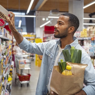 Mid adult multiracial male reaching for cereal box on a shelf in a grocery store aisle. He is holding a paper bag filled with groceries, including a bottle and vegetables. The aisle is brightly lit and organized with various products on display.