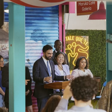 Mayor Mamdani and a group of people stand at a podium giving a speech in the colorful indoor space of La Marqueta with hanging lights and a crowd watching. The atmosphere is lively and festive, with blue and pink decor and a sign reading “Maruka Café Galería."