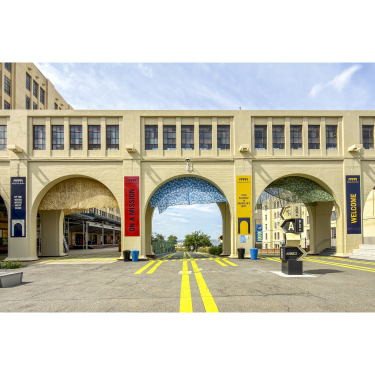 A wide archway with beige walls and colorful banners reading ON A MISSION and STAY INSPIRED. Yellow road lines lead under the arches, with trees and blue sky visible beyond.