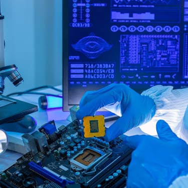 A person in lab gloves installs a computer chip onto a motherboard beside a microscope, with a monitor displaying blue technical diagrams in the background.