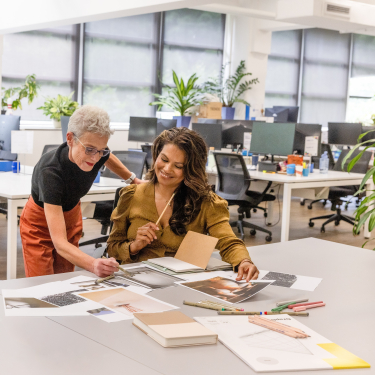 Two women collaborate at a desk, looking at printed images and drawings.
