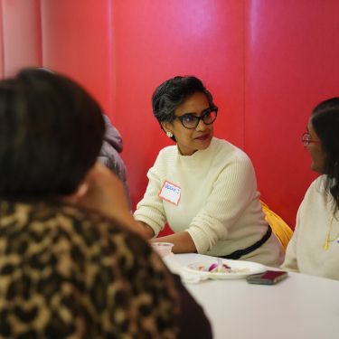 Three women sit around a table at a meeting, with two women wearing name tags facing each other while talking.