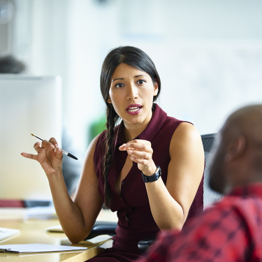 A woman in a sleeveless maroon top gesturing while speaking to a colleague in an office.