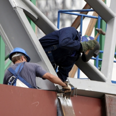 Two construction workers in blue uniforms and helmets are assembling a large metal structure. One worker is using a tool while the other supports the structure. They are on a construction site with green and brown structural elements in the background. Two construction workers in blue uniforms and helmets are assembling a large metal structure. One worker is using a tool while the other supports the structure. They are on a construction site with green and brown structural elements in the background.