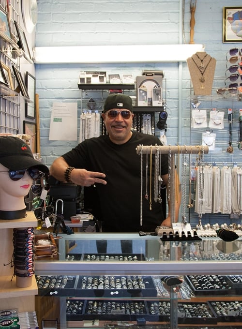 A man wearing sunglasses and a black cap stands behind the counter of a jewelry store, smiling and making a peace sign. The shop is filled with necklaces, rings, sunglasses, and accessories on display.
