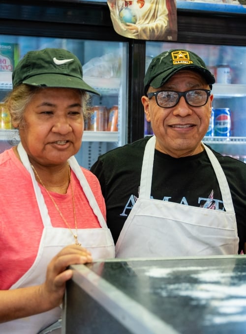 Two smiling people wearing aprons and hats stand behind a counter in a kitchen or diner. One gives a thumbs up. Shelves with food and a coffee container are visible in the background.