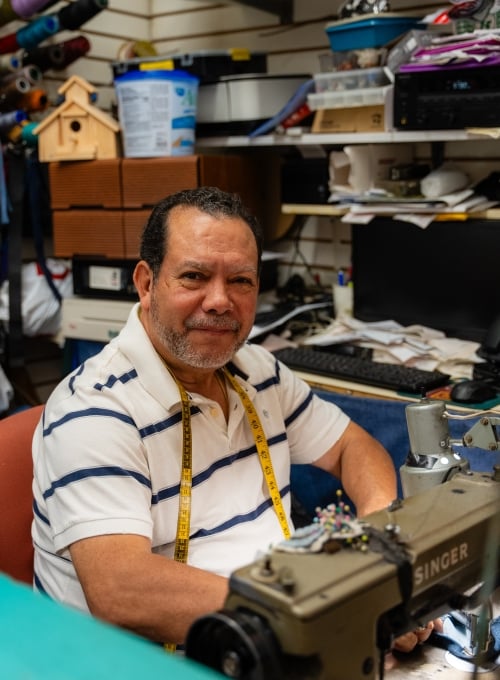 A man with short hair and a beard sits at a sewing machine in a workshop, smiling. He wears a striped shirt and a yellow tape measure around his neck. Shelves with various supplies and tools are in the background.