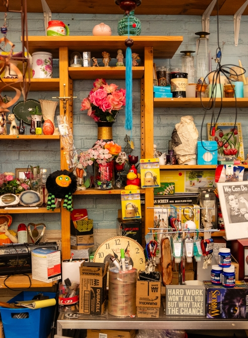 A cluttered thrift store shelf is filled with various items, including clocks, flowers, figurines, books, mugs, toys, and colorful decorations, creating a busy and eclectic display against a brick wall.