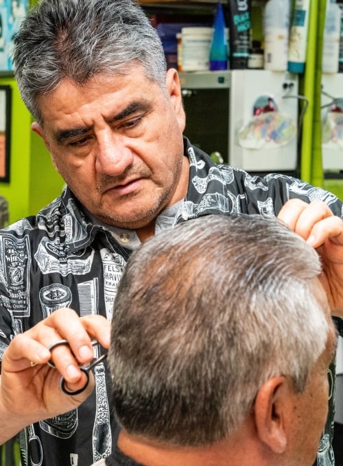 A barber with gray hair is cutting a clients hair using scissors. The client has short, gray hair. The barber is focused on his work inside a shop, with various hair products visible in the background.