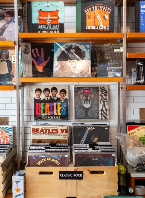 A display shelf holds an assortment of vinyl records. The top shelf features albums from bands and jazz artists. Below, a crate labeled Classic Rock contains various rock albums, including those by The Beatles, arranged in an organized manner.