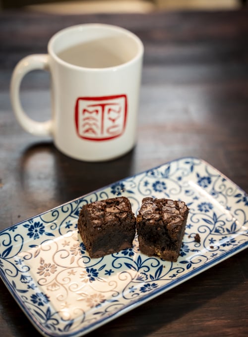 Two brownies on a blue and white floral-patterned plate, placed on a dark wooden table. A white mug with a red design is in the background.