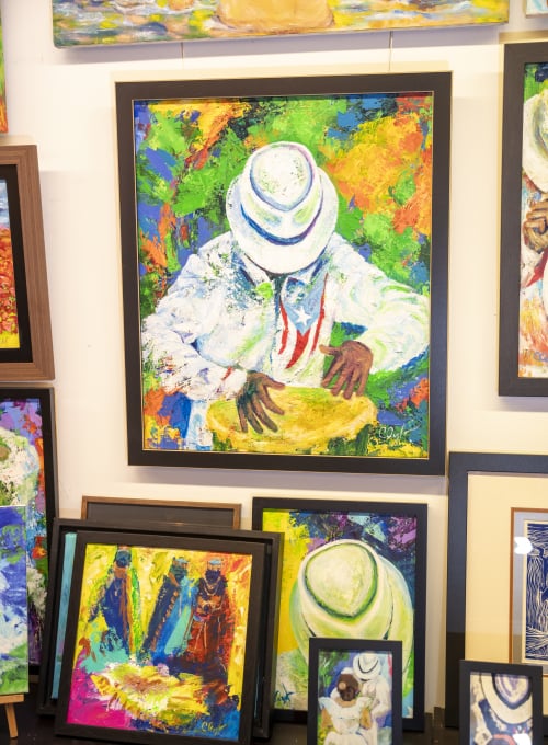 A vibrant display of colorful paintings, including a large framed painting of a figure in a white hat playing a drum. Other smaller paintings featuring abstract and colorful scenes are arranged around it.