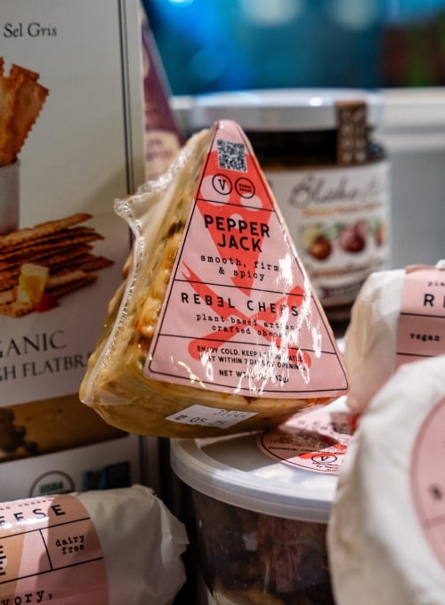 A wedge of Pepper Jack plant-based cheese with a pink label sits among various other food items. The label mentions smooth, firm &amp;amp; spicy and dairy free. Other labeled plant-based cheeses are partially visible around it.