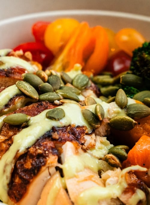 A close-up of a bowl with sliced roasted chicken topped with pumpkin seeds and drizzled with a light green sauce. Accompanied by vibrant cherry tomatoes and broccoli florets, creating a colorful and appetizing presentation.