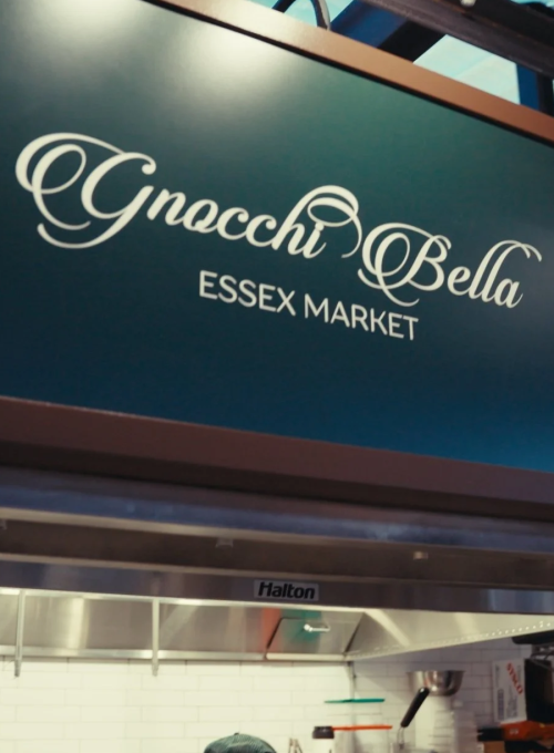 A sign reads Gnocchi Bella ESSEX MARKET above a kitchen area with a stainless steel backsplash and utensils hanging on the wall.