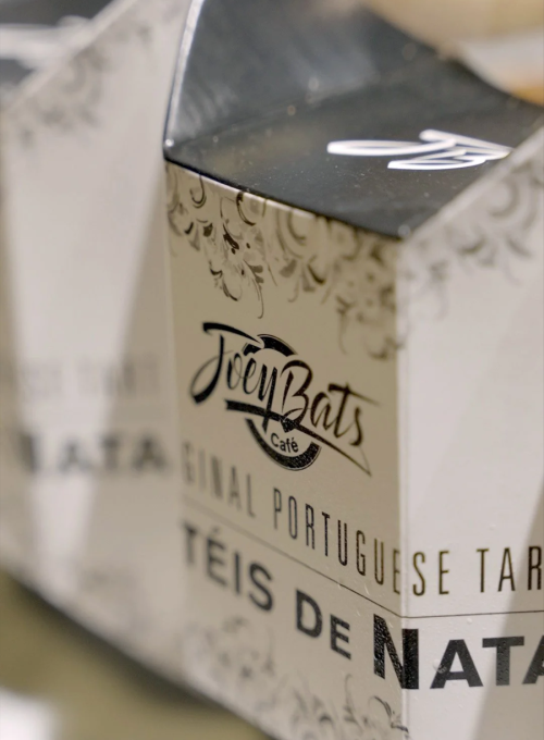 Close-up of a box with the logo Joey Bats Café and text reading Original Portuguese Tarts and Pães de Nata printed on the packaging in decorative fonts.