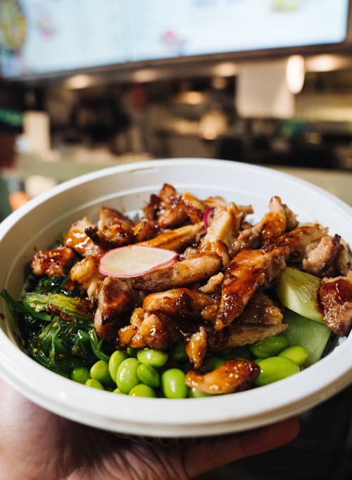 A hand holds a bowl filled with glazed grilled chicken, sliced radish, edamame, seaweed salad, and greens, with a blurred restaurant counter in the background.