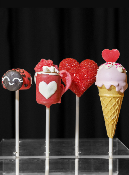 Four decorative cake pops on sticks: a smiling round pop with mustache, a red mug with a heart and marshmallows, a red glittery heart, and a pink ice cream cone with sprinkles, all against a black background.